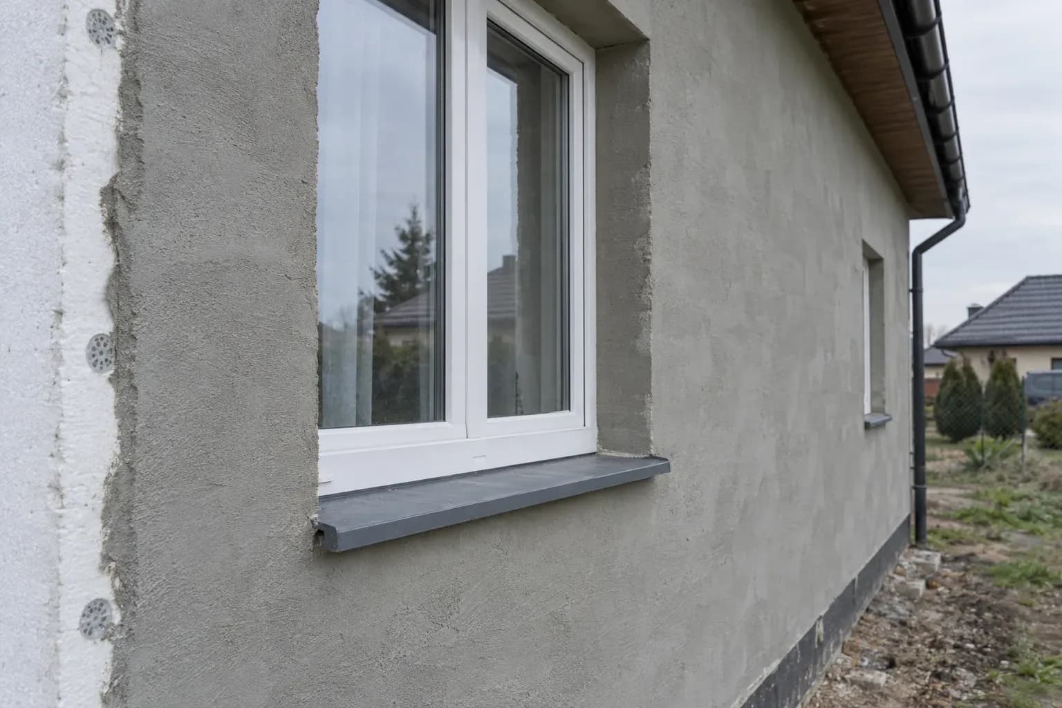 Narożnik ściany z warstwą ocieplenia i tynkiem podkładowym, obok okno z białą ramą